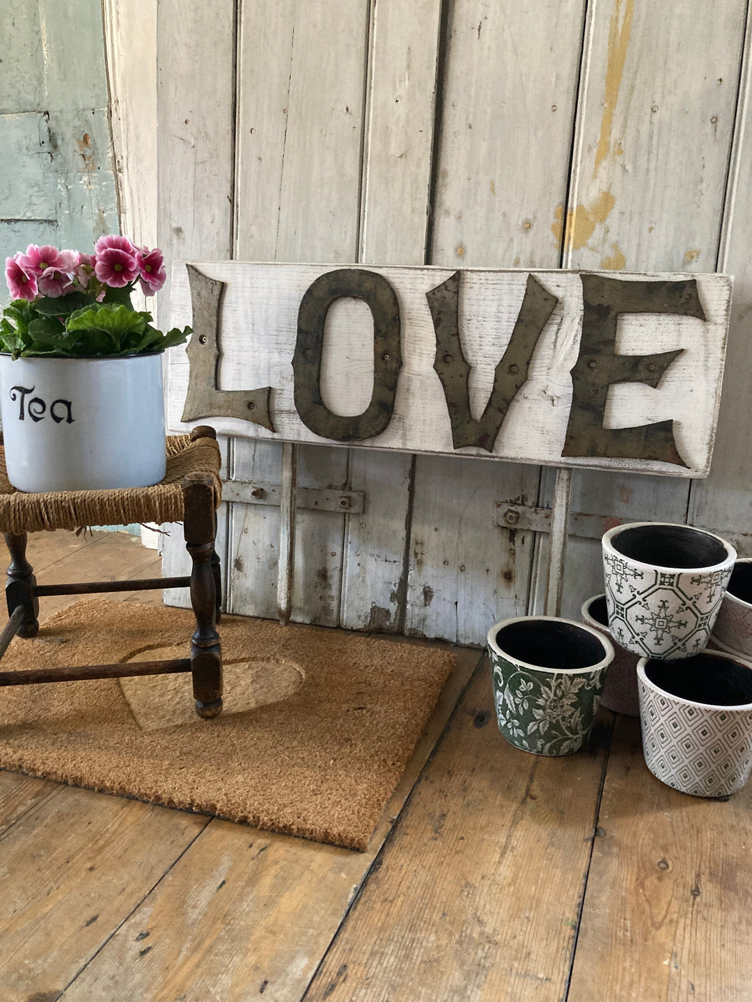 Vintage metal letters spelling LOVE on a handmade wooden sign, surrounded by flowerpots and an old kitchen canister with flowers