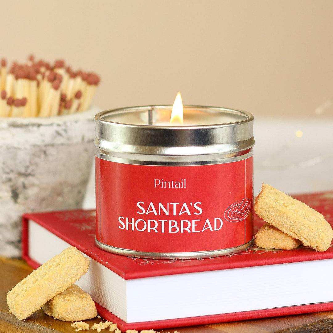 Pintail Candles Santa's Shortbread Classic Tin Candle lit on a book with shortbread biscuits nearby.