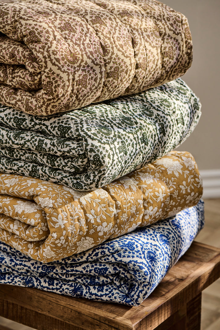 Stack of floral quilted throws in brown, green, yellow, and blue on a wooden bench. Cozy home decor from Source for the Goose, Devon.