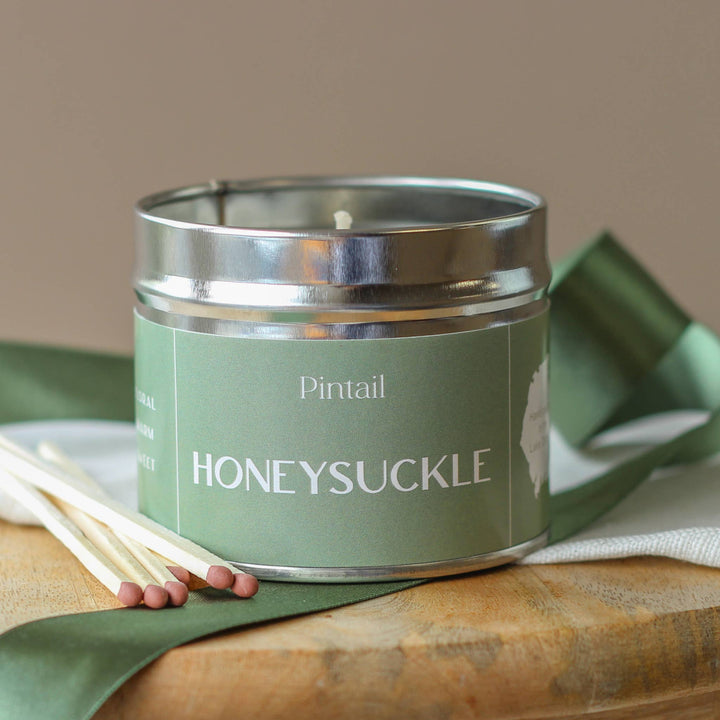 Pintail Honeysuckle Classic Tin Candle beside matchsticks on a wooden tabletop, evoking a fresh, floral ambiance.