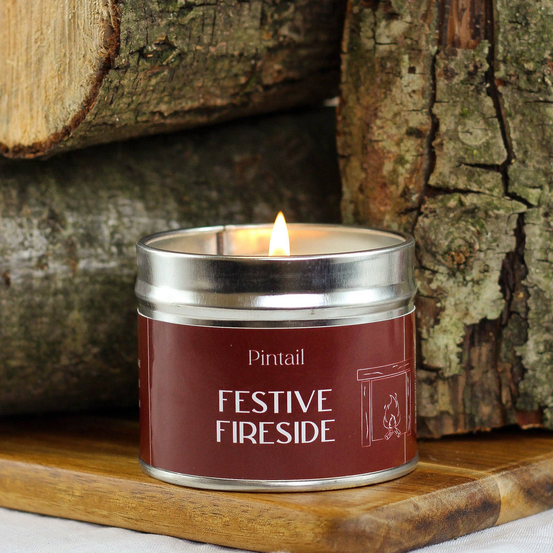 Festive Fireside Classic Tin Candle by Pintail, burning brightly beside stacked logs, handmade in Cumbria, available at Source for the Goose in Devon.