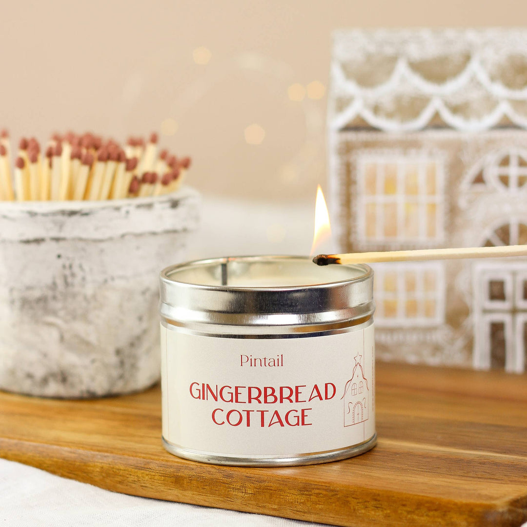 Pintail Candles Gingerbread Cottage Classic Tin Candle being lit on a wooden surface, surrounded by cozy decor.