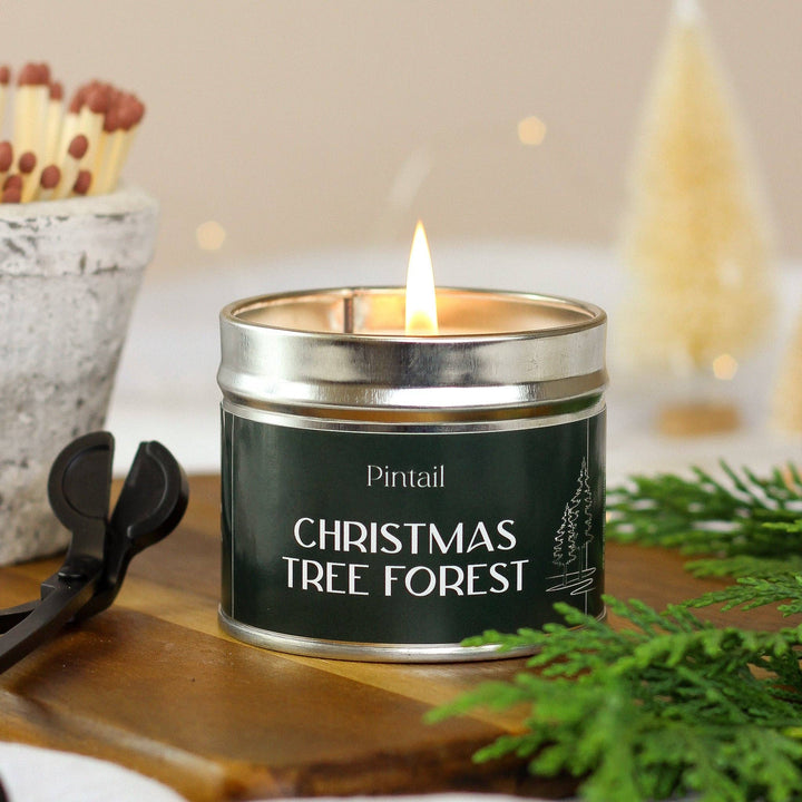 Pintail Candles Christmas Tree Forest Classic Tin Candle, hand-poured in Cumbria, next to greenery and matchsticks on a wooden surface