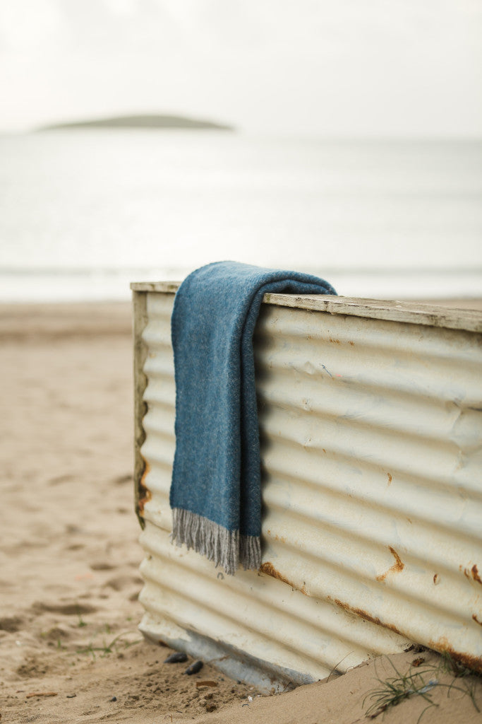 Tweedmill Lifestyle Boa Pure New Wool Blanket in Inky Blue with grey tassels on the beach, Source for the Goose Devon.