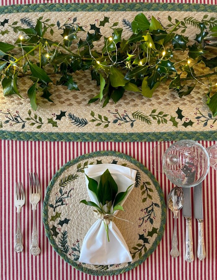 Jute Christmas placemat with holly design from Source for the Goose Devon on red-striped table setting with silverware and greenery.