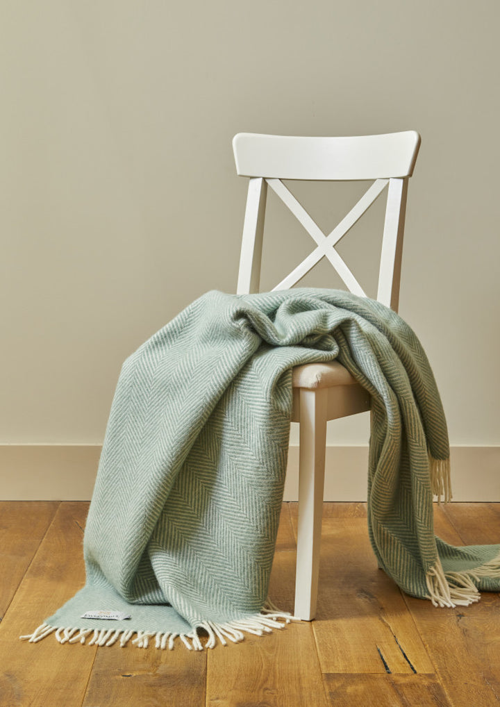 Sea green Tweedmill Fishbone Blanket with cream tassels draped over a white chair on a wooden floor, cozy home decor from Source for the Goose Devon.