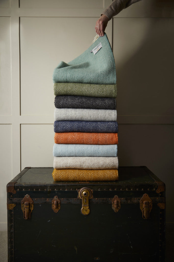 Stack of Tweedmill Fishbone Blankets in various colors on a vintage trunk, adding cosiness and style to any living space from Source for the Goose Devon.