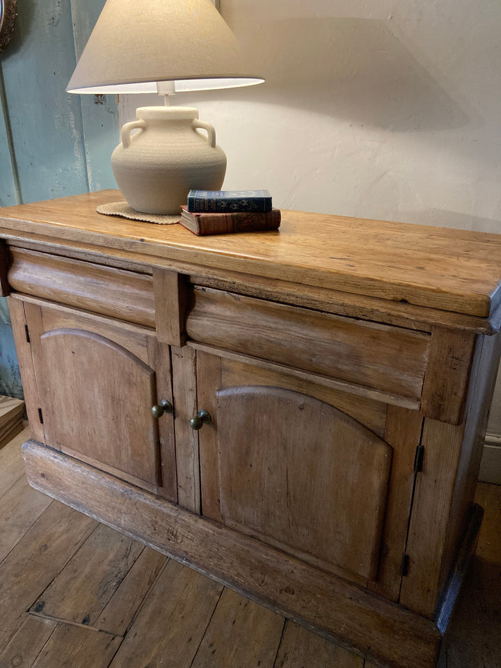 Rustic antique pine sideboard from Source for the Goose Devon, featuring two drawers and brass handles, styled with lamp.