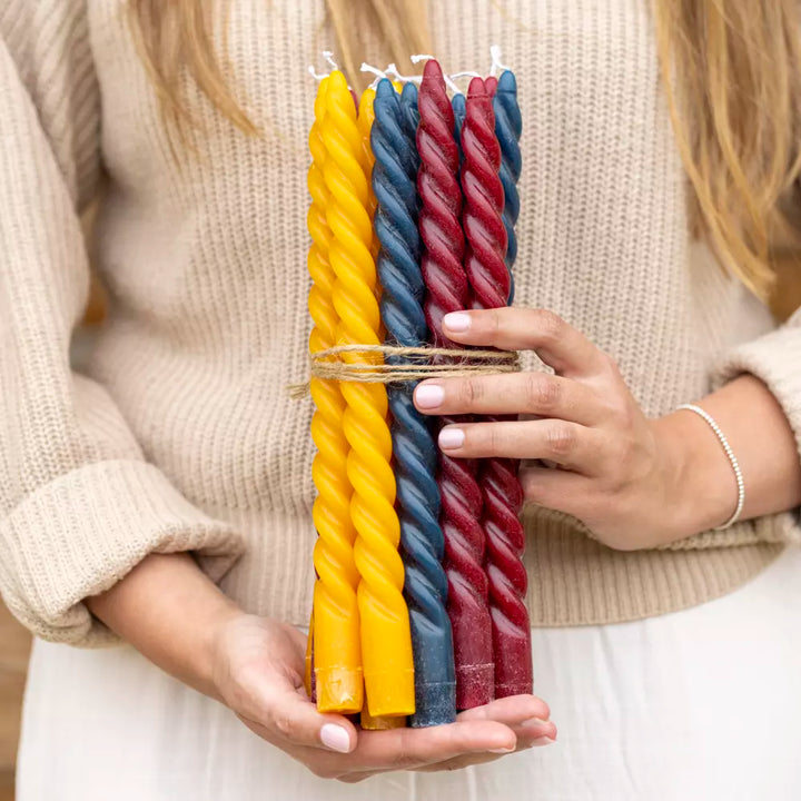 Set of four twisted ochre, blue, and burgundy dinner candles held by a person, showcasing elegance for dining occasions.