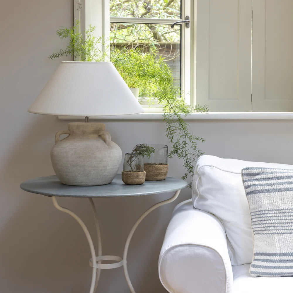 Stoneware Amphora Lamp with cream shade on side table near sofa in light-filled room with plants in the background. Source for the Goose Devon.