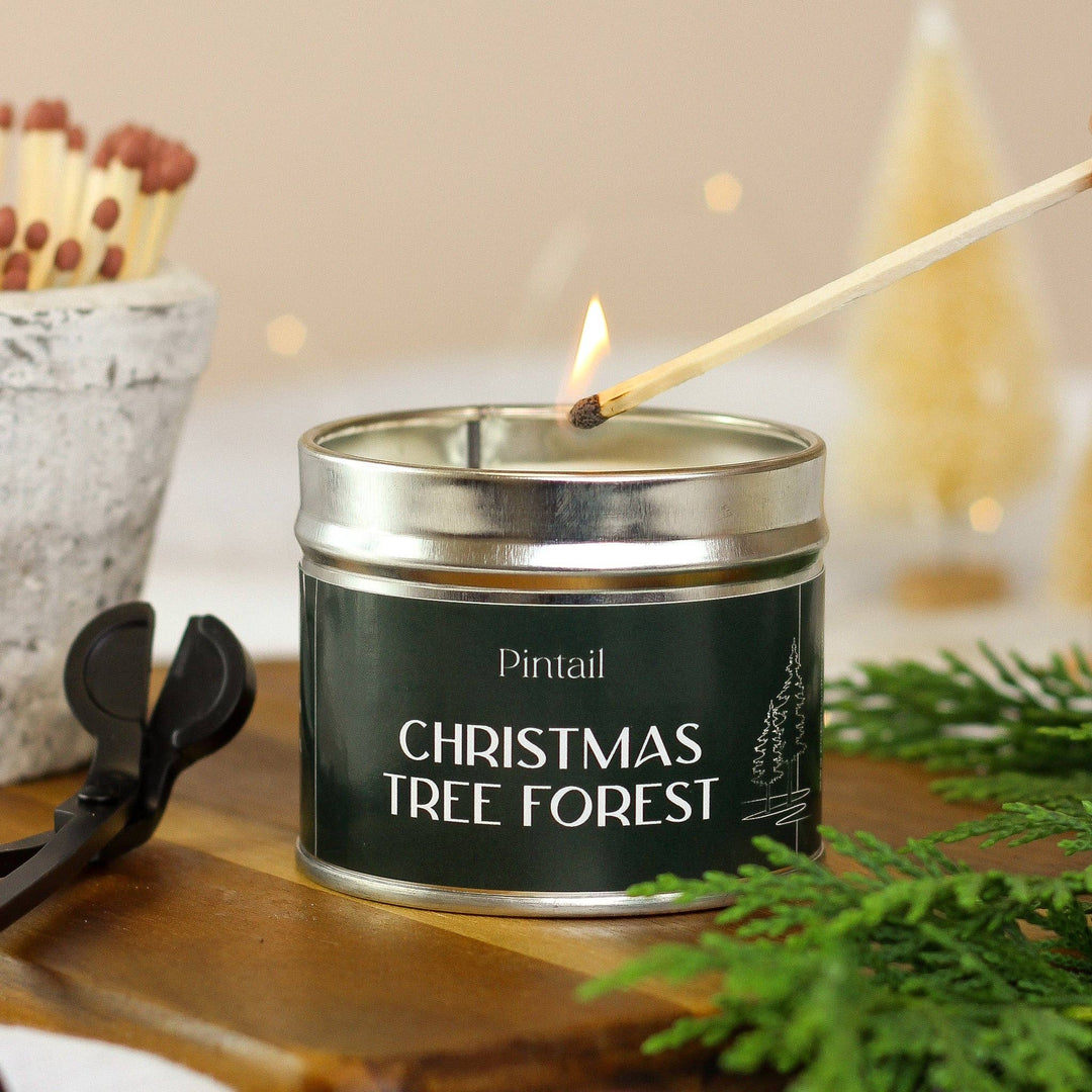 Pintail Candles Christmas Tree Forest Classic Tin Candle being lit with matches on a wooden table surrounded by festive decor.