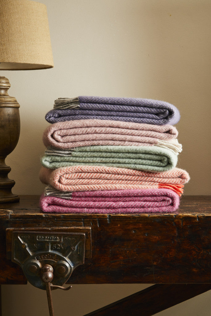 Stack of Tweedmill herringbone wool blankets in various colors on a rustic wooden table with a lamp in the background, available from Source for the Goose, Devon