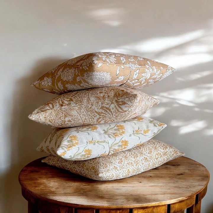 Stack of four decorative cushions on a wooden table, featuring vintage-inspired patterns in soft hues.