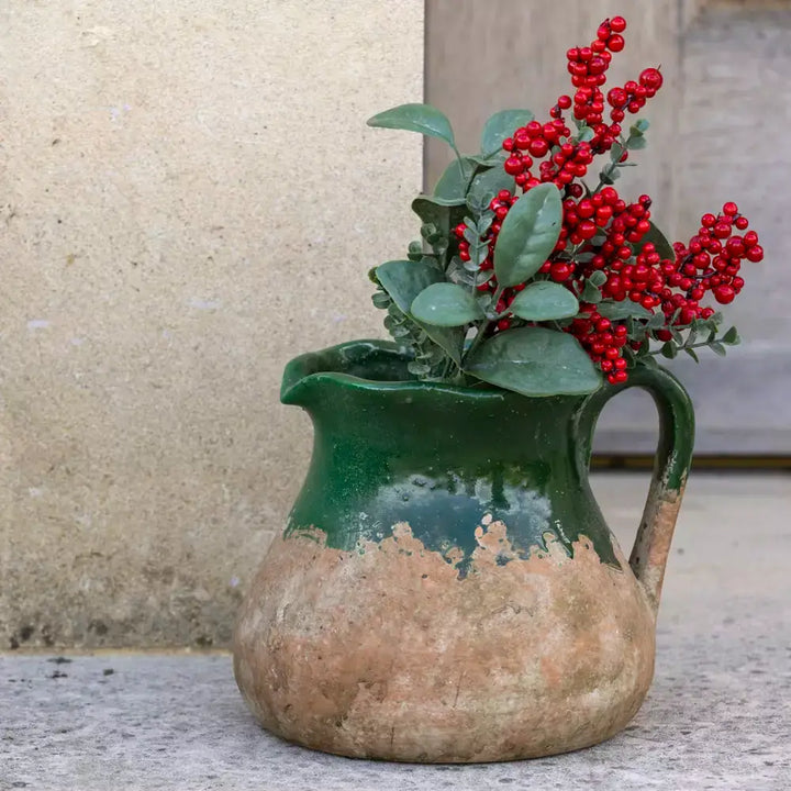 Vintage rustic green terracotta jug with red berries and green leaves, showcasing earthy charm and traditional design with a sturdy handle and spout.