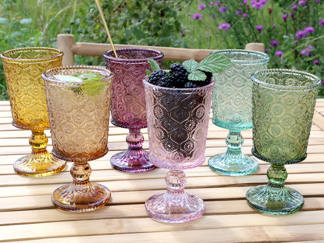 Set of six vintage style coloured wine glasses by Source for the Goose, Devon, arranged on a wooden table with a floral background.