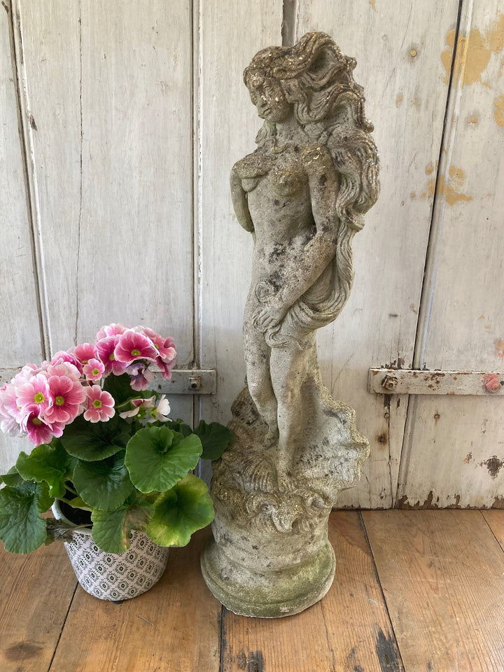 Stone garden statue of a lady with wavy hair and shell base, beside pink flowers. Source for the Goose, Devon.