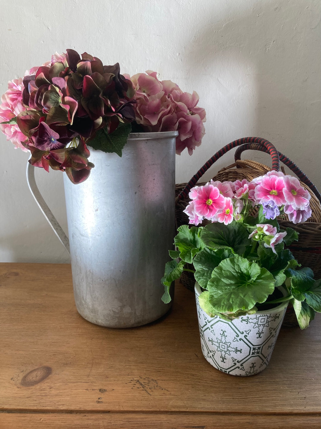 Vintage tall aluminium jug filled with flowers beside a potted plant on a wooden surface.