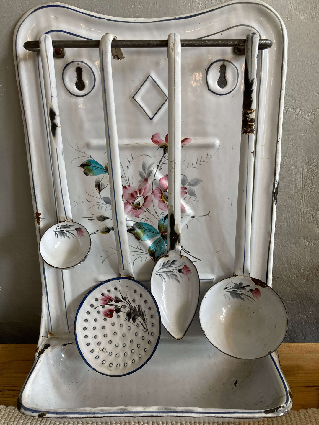 Vintage French enamel spoon rest with blue and pink floral design, featuring rustic utensils hanging on a white distressed enamel panel.