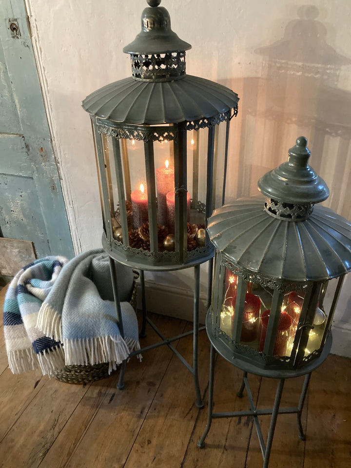 Vintage grey lanterns on stands with candles, beside a cozy blanket, adding warmth and elegance to a room. Source for the Goose Devon.